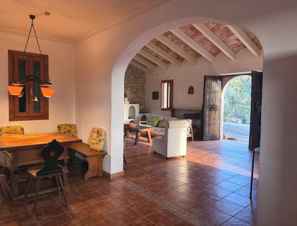 Rustic Mediterranean living space with arched doorway, terracotta tile floor, wooden dining table, stone wall and open door to the garden.