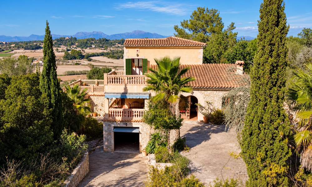 Mediterranean stone villa with terracotta roof, balcony, palm trees and cypress trees overlooking rural farmland and distant mountains.