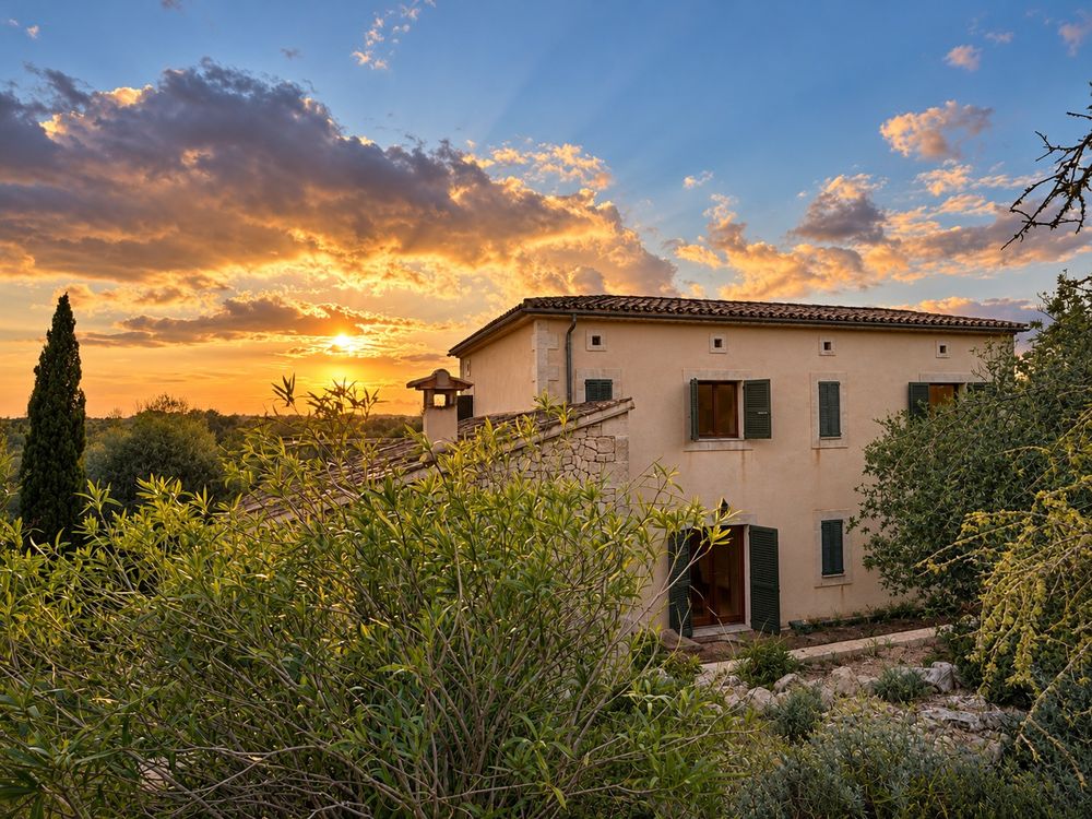 Mediterranean stone villa with green shutters surrounded by lush greenery and cypress trees under a vivid orange and purple sunset sky.
