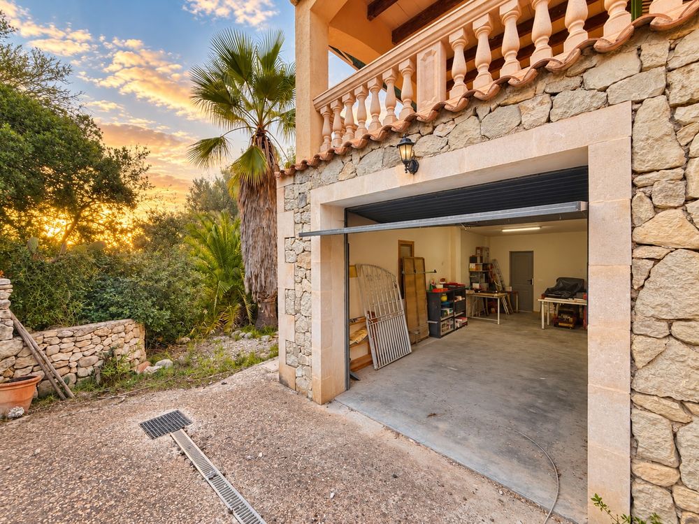 Stone villa garage with open door and balcony above, framed by palm trees and Mediterranean greenery at a golden sunset.