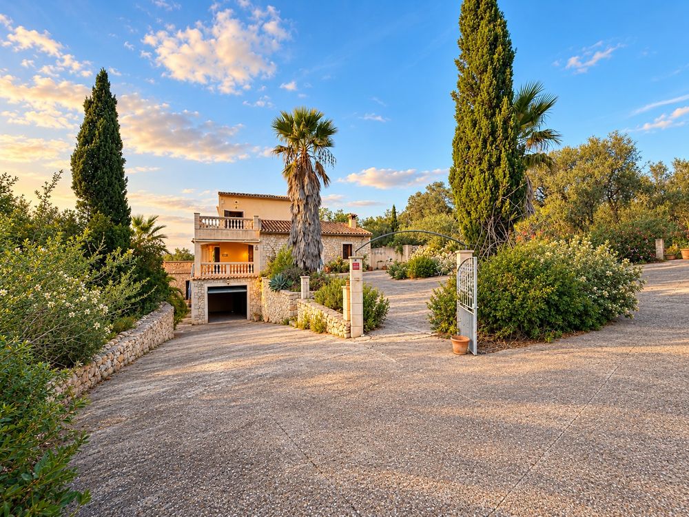 Mediterranean stone villa with balcony, framed by tall palm and cypress trees, reached by a wide gravel driveway under a blue sky.