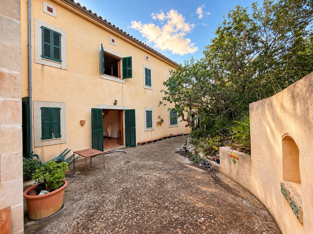 Charming two-story Mediterranean stone house with green shutters, open wooden doors, a gravel courtyard, potted plants and lush trees.