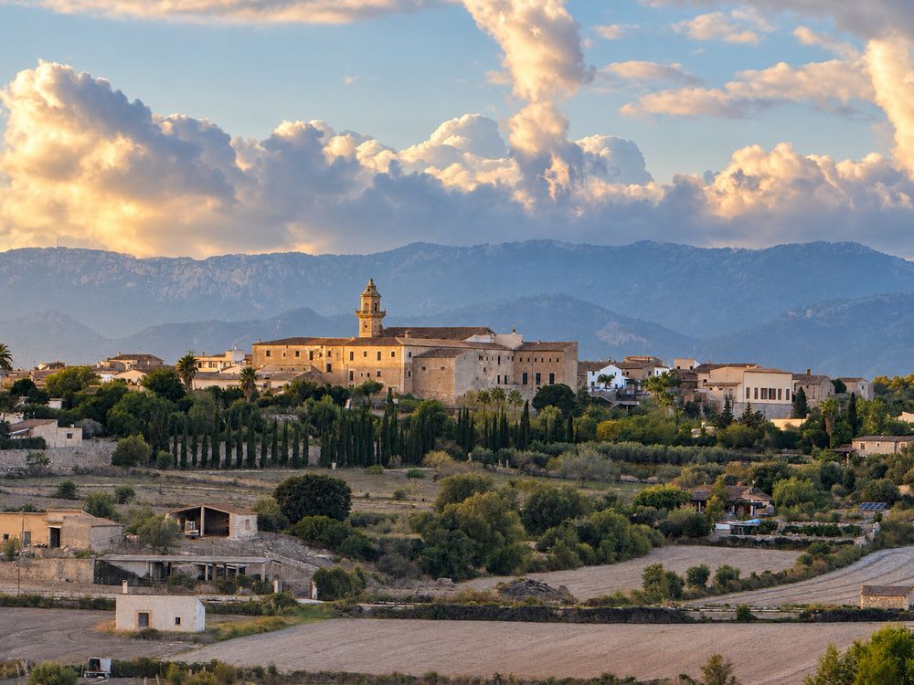 Hilltop village with a stone church tower overlooking green farmland and cypress trees, with misty mountains and dramatic clouds behind.