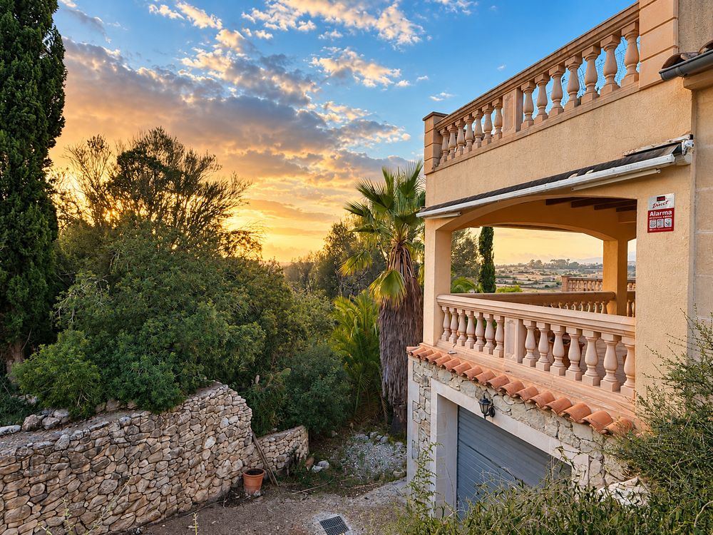 Mediterranean villa with terracotta-tiled balcony and garage overlooking palm trees and a stone wall at a colorful golden sunset.