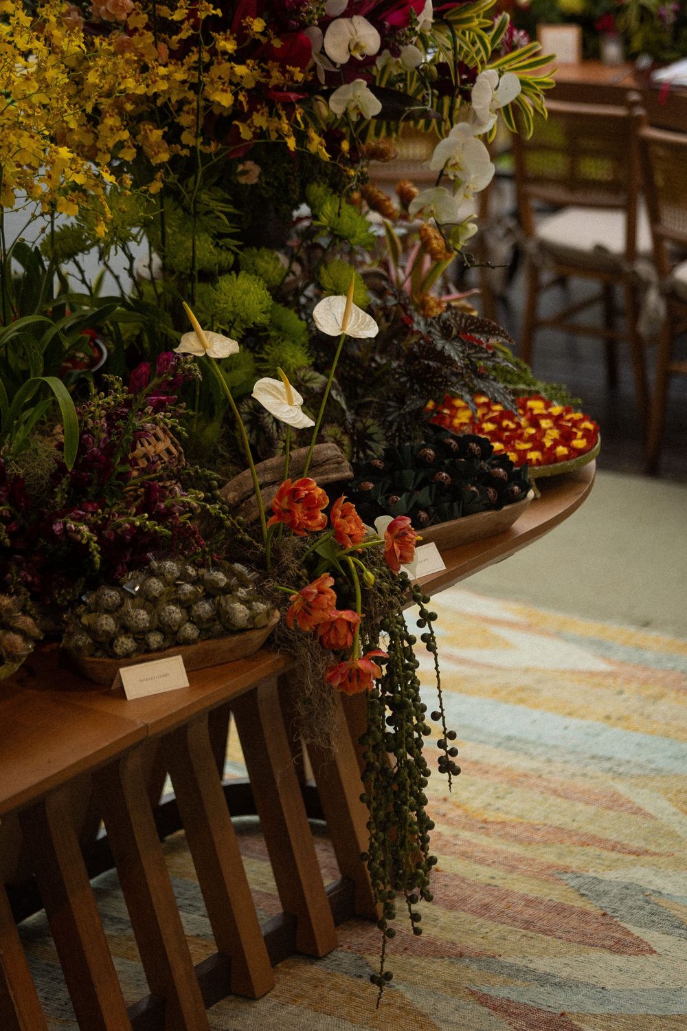 Wooden display table with white orchids, orange blooms, trailing greenery, and bowls of berries and fruit beside a patterned rug.