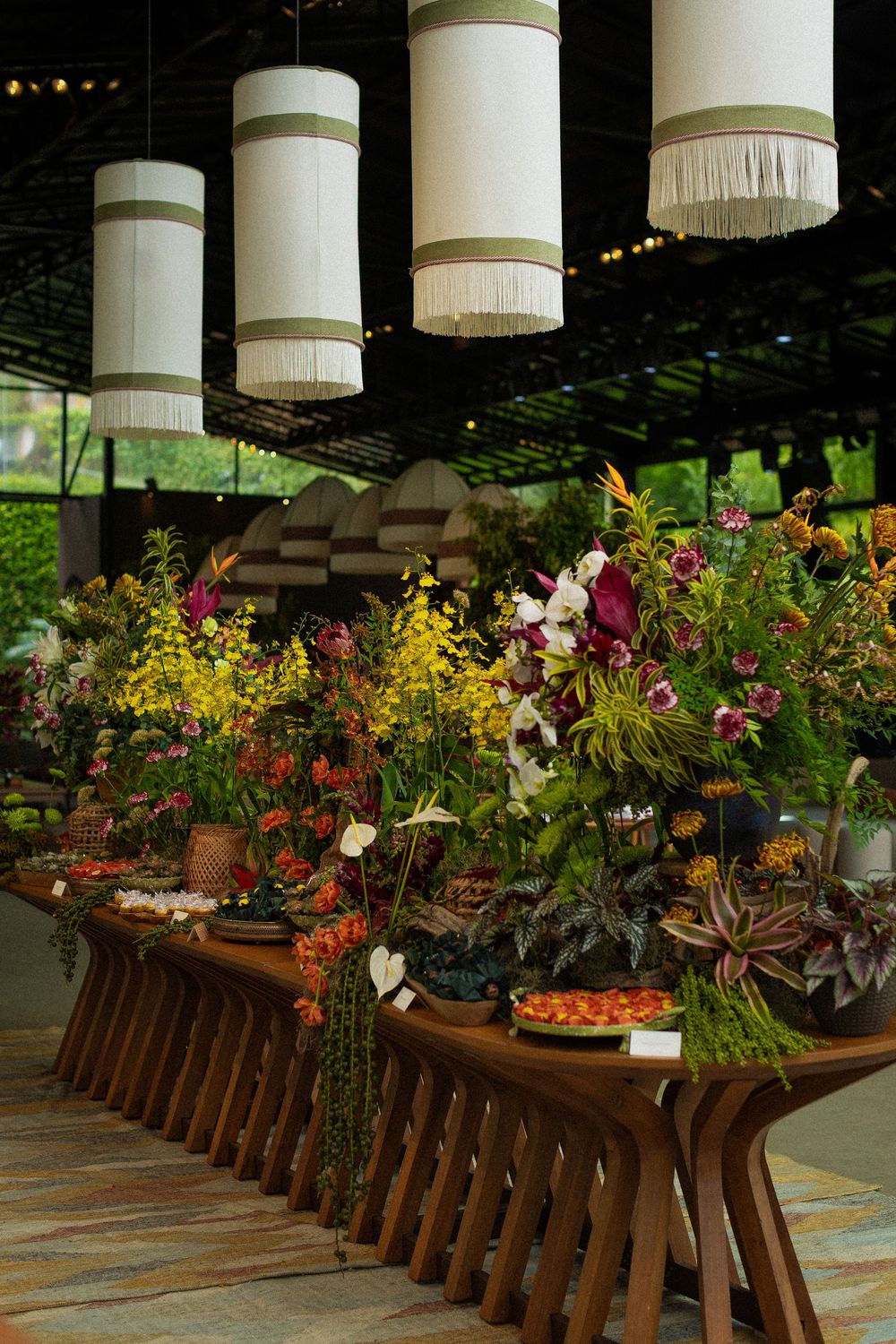 Colorful floral arrangement of yellow, orange and pink blooms on a curved wooden table beneath white fringed cylindrical pendant lamps.