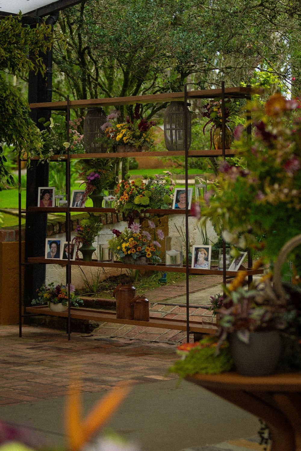 Rustic metal and wood shelving displays framed photos and colorful flower arrangements in a lush garden memorial setting.