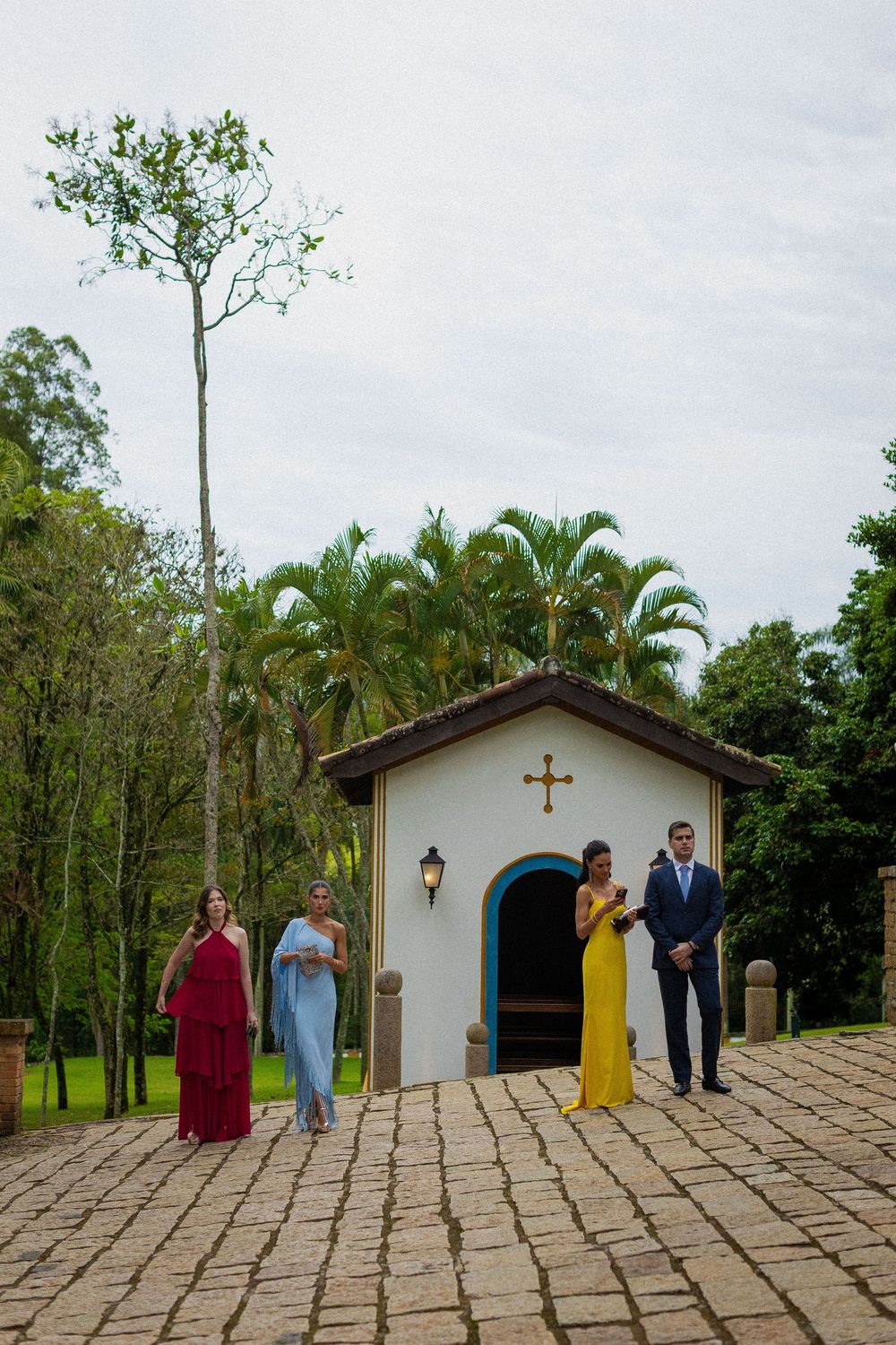 Wedding guests in red, blue and yellow dresses and a man in a navy suit stand outside a small white chapel on a cobblestone path.