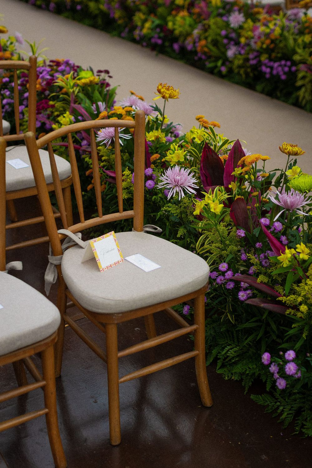 Wooden chiavari chairs with cream seat cushions and small place cards beside a colorful floral border of asters, zinnias, and purple blooms.