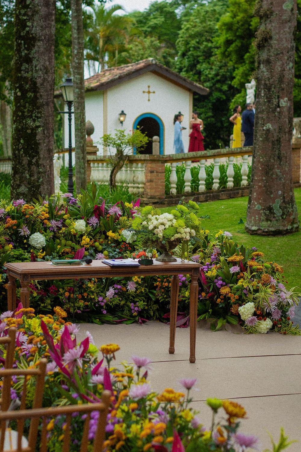 Wooden table surrounded by colorful flower arrangements at an outdoor garden wedding with a small white chapel and guests in the background.
