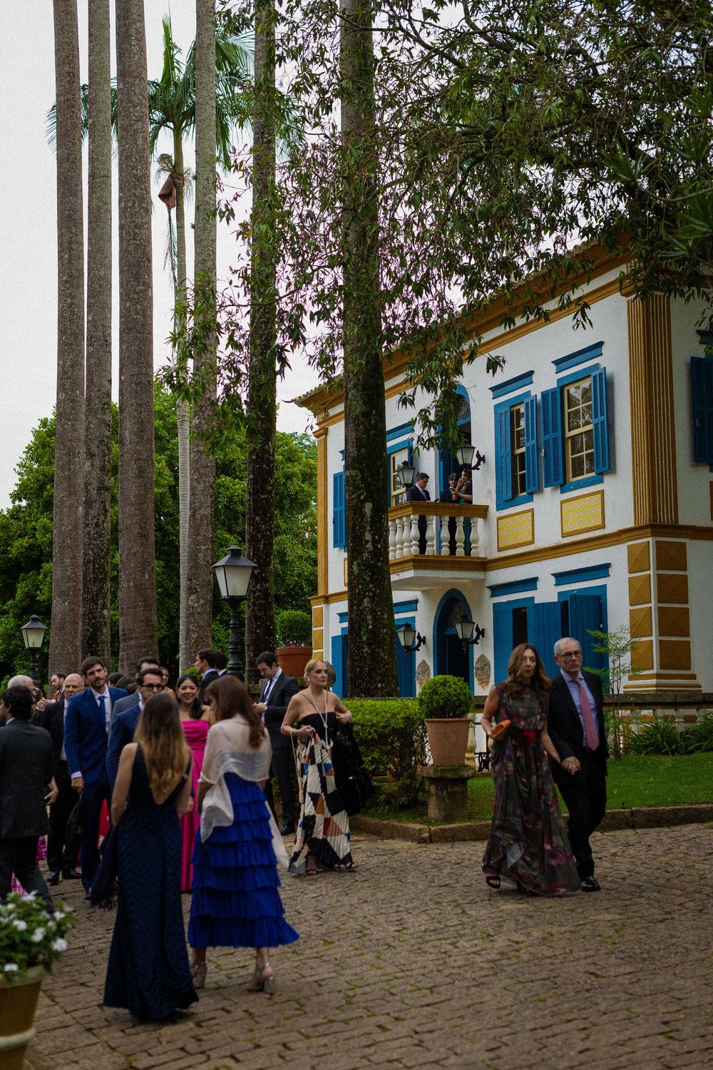 Elegantly dressed wedding guests gather on a cobblestone courtyard beside a blue and white colonial mansion surrounded by tall palm trees.