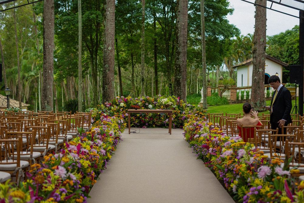 Outdoor wedding ceremony aisle lined with colorful flower arrangements and wooden chairs among tall trees, with a man in a suit nearby.