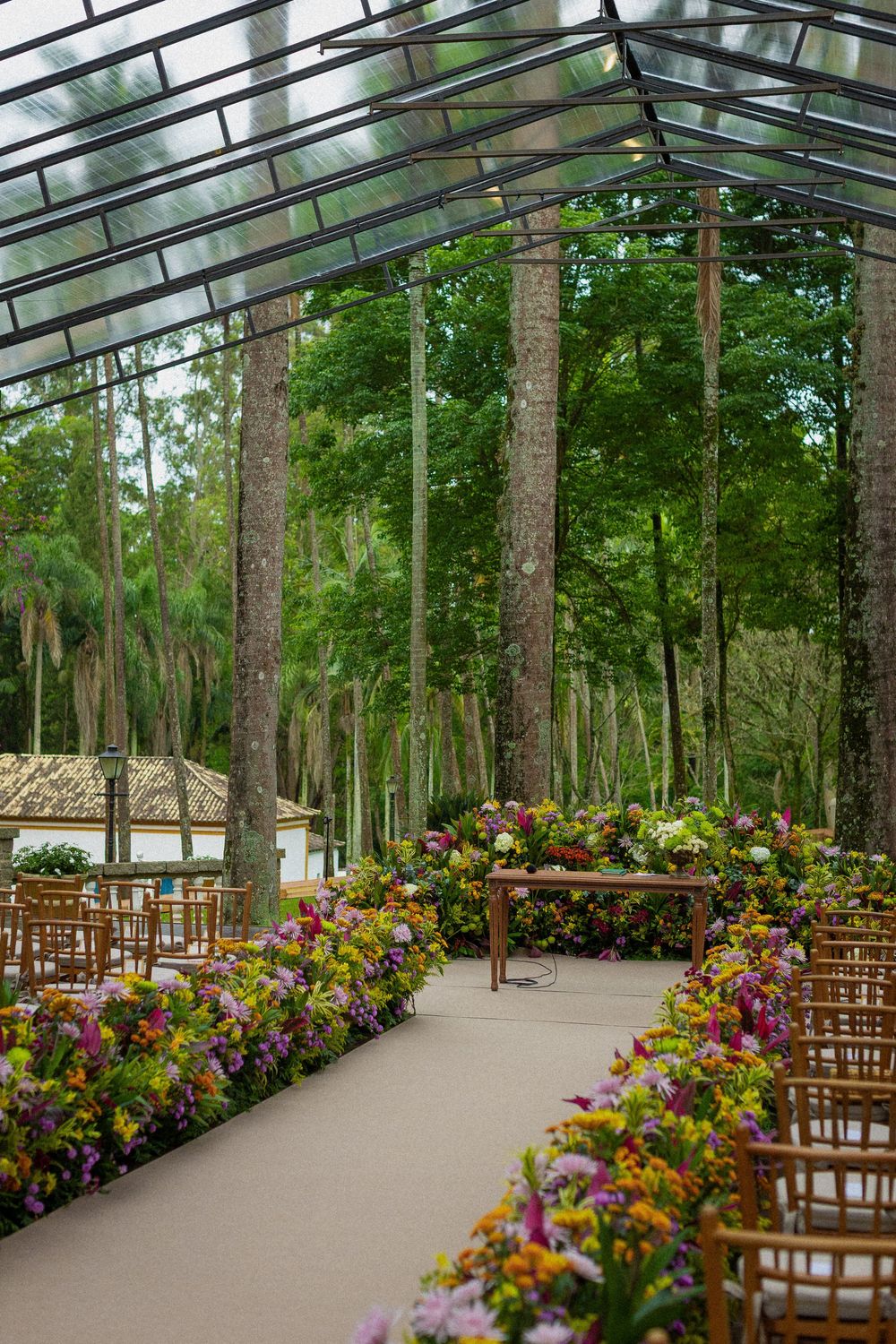 Outdoor wedding ceremony aisle lined with colorful flower arrangements and wooden chairs beneath a glass roof among tall forest trees.