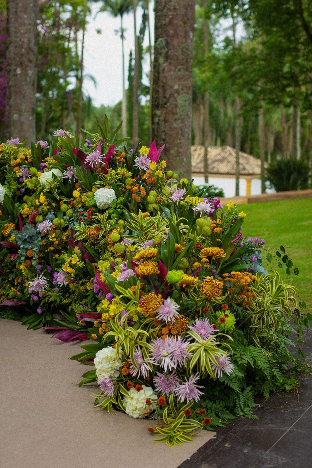 Lush floral arrangement of purple, orange, yellow and white blooms with green foliage along a garden path, with tall trees and lawn behind.