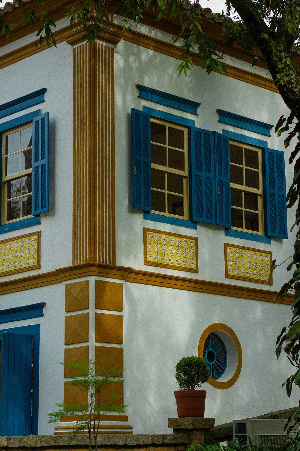 Colonial-style corner building with white walls, blue shutters, golden ornamental panels, a round window and a potted topiary shrub.