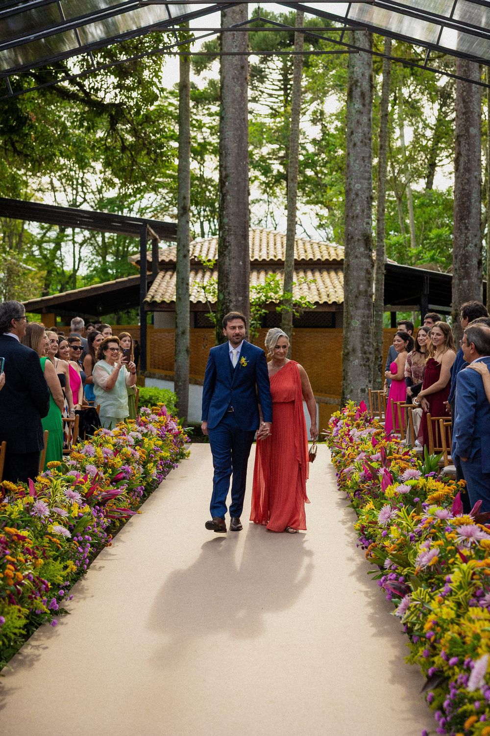 Groom in a navy suit walks hand in hand with a woman in a coral dress down an aisle lined with colorful flowers at an outdoor wedding.