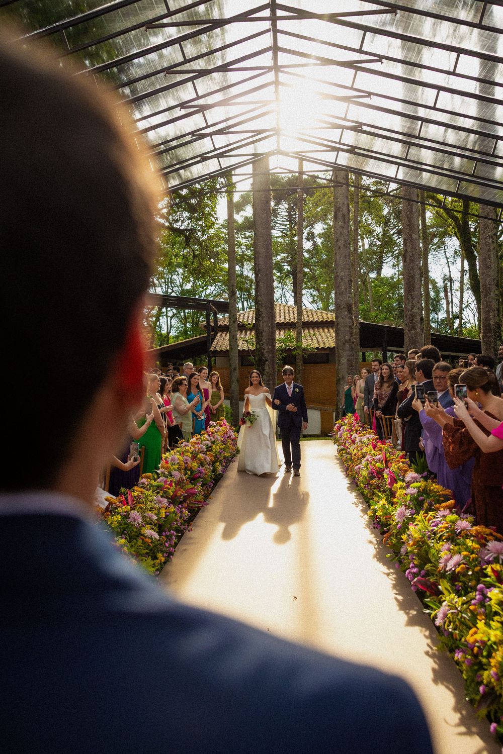 Bride and groom walk down a flower-lined aisle through a sunlit glass-roofed garden venue as wedding guests watch and take photos.