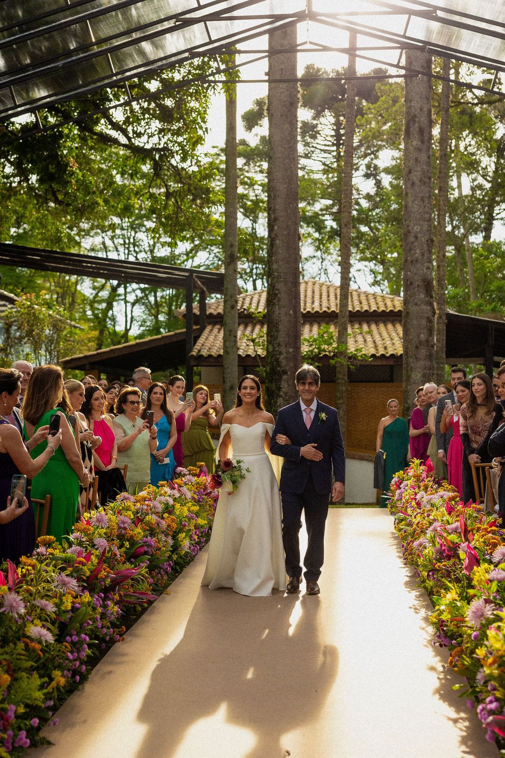 Bride in a white gown walks down an aisle lined with colorful flowers beside a man in a navy suit as guests watch at an outdoor wedding.