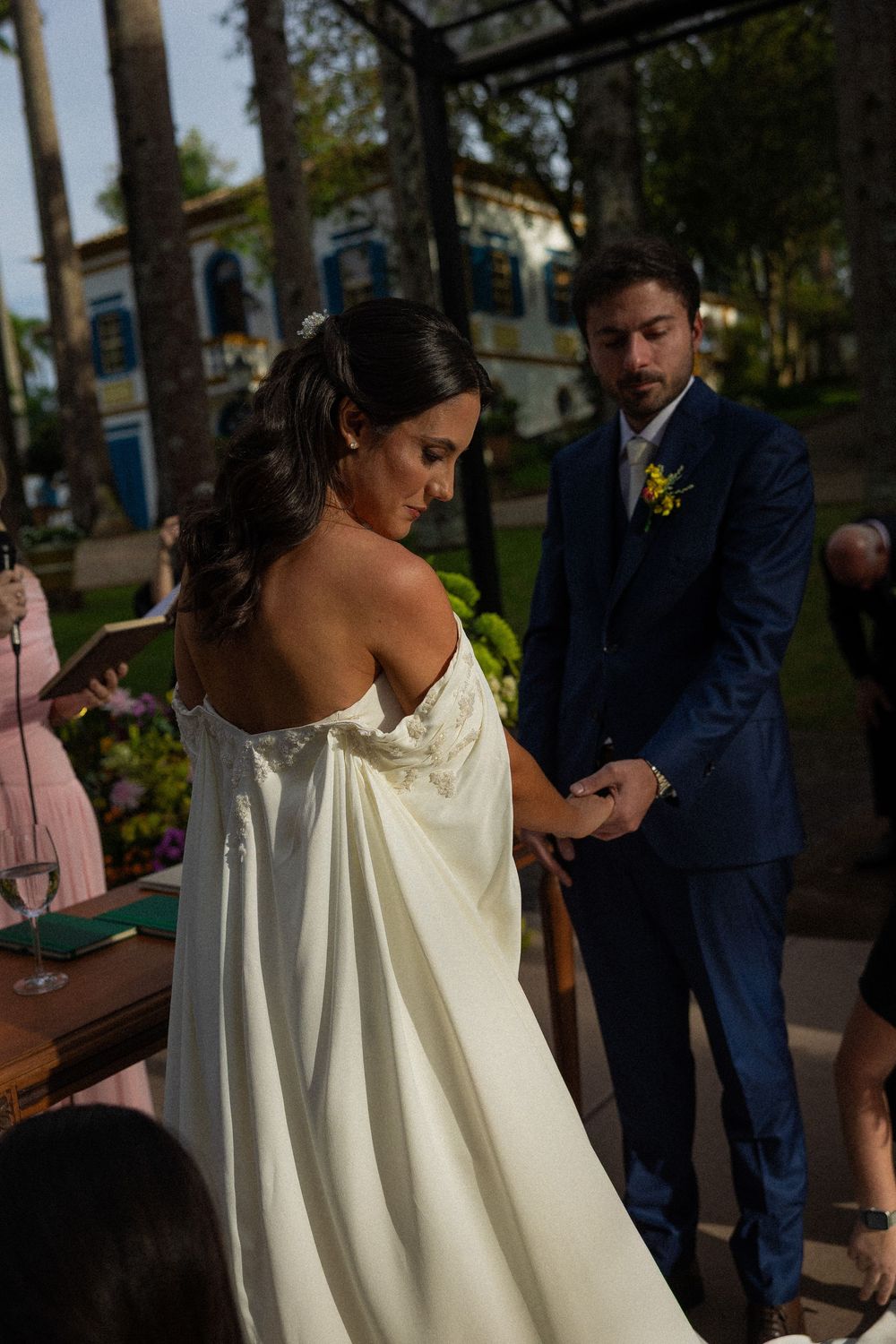 Bride in an off-shoulder white gown holds hands with a groom in a navy suit during an outdoor wedding ceremony under trees.