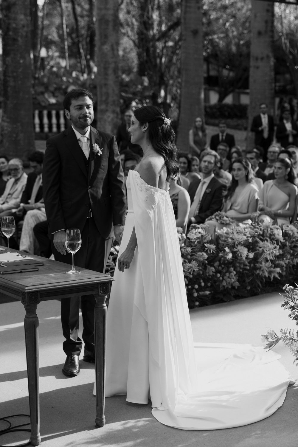 Bride in a flowing white gown with cape sleeves stands beside the groom at an outdoor wedding ceremony as guests watch among lush flowers.