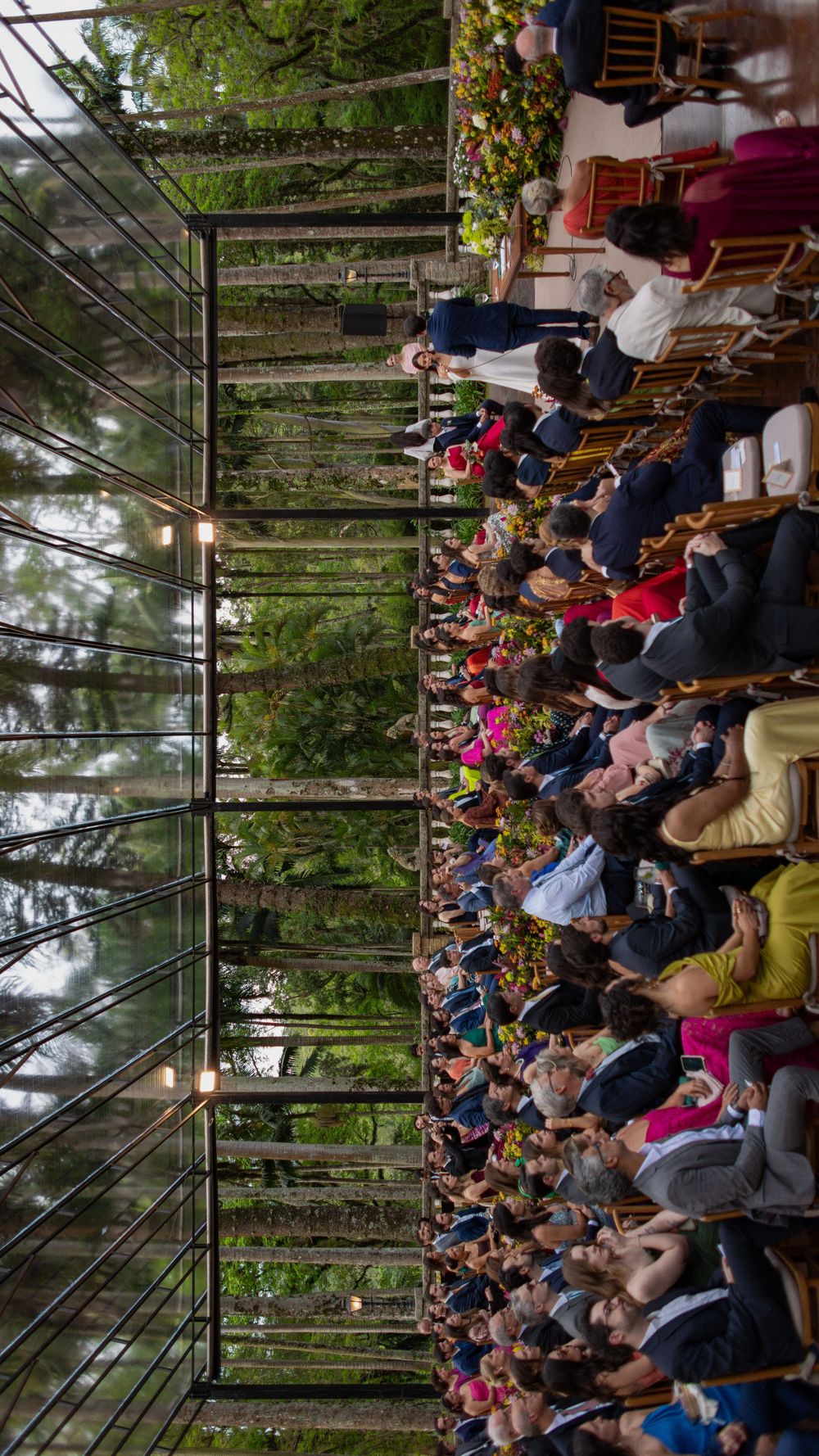 Wedding guests seated in rows of wooden chairs during an outdoor ceremony inside a glass pavilion surrounded by tall forest trees.