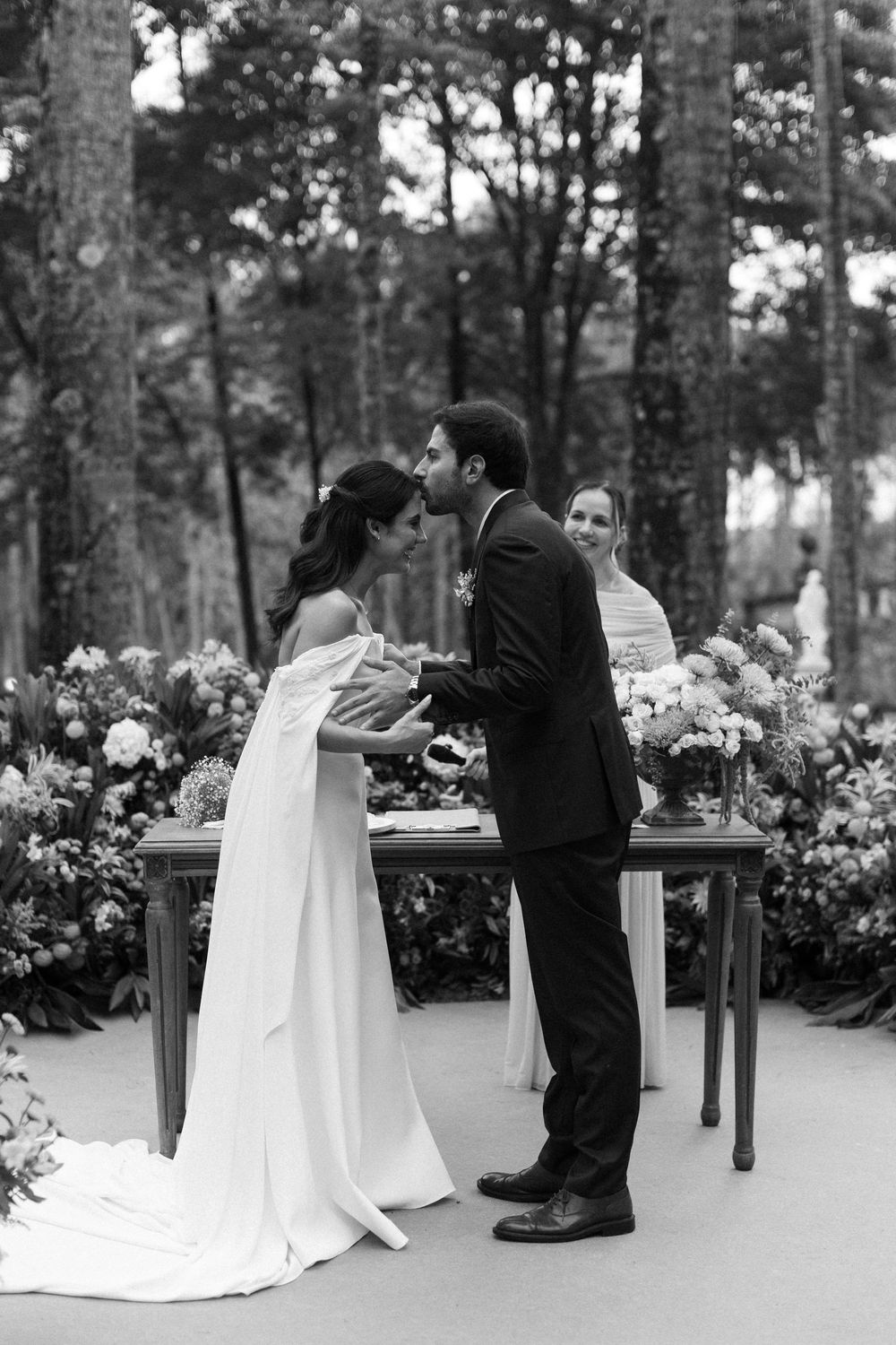 Black and white photo of a bride and groom sharing a kiss at an outdoor forest wedding ceremony surrounded by flowers and an officiant.