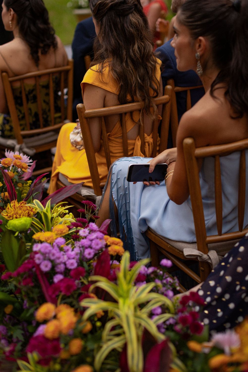 Wedding guests in yellow and blue dresses sit on wooden chairs behind a colorful floral arrangement of pink, orange and purple blooms.