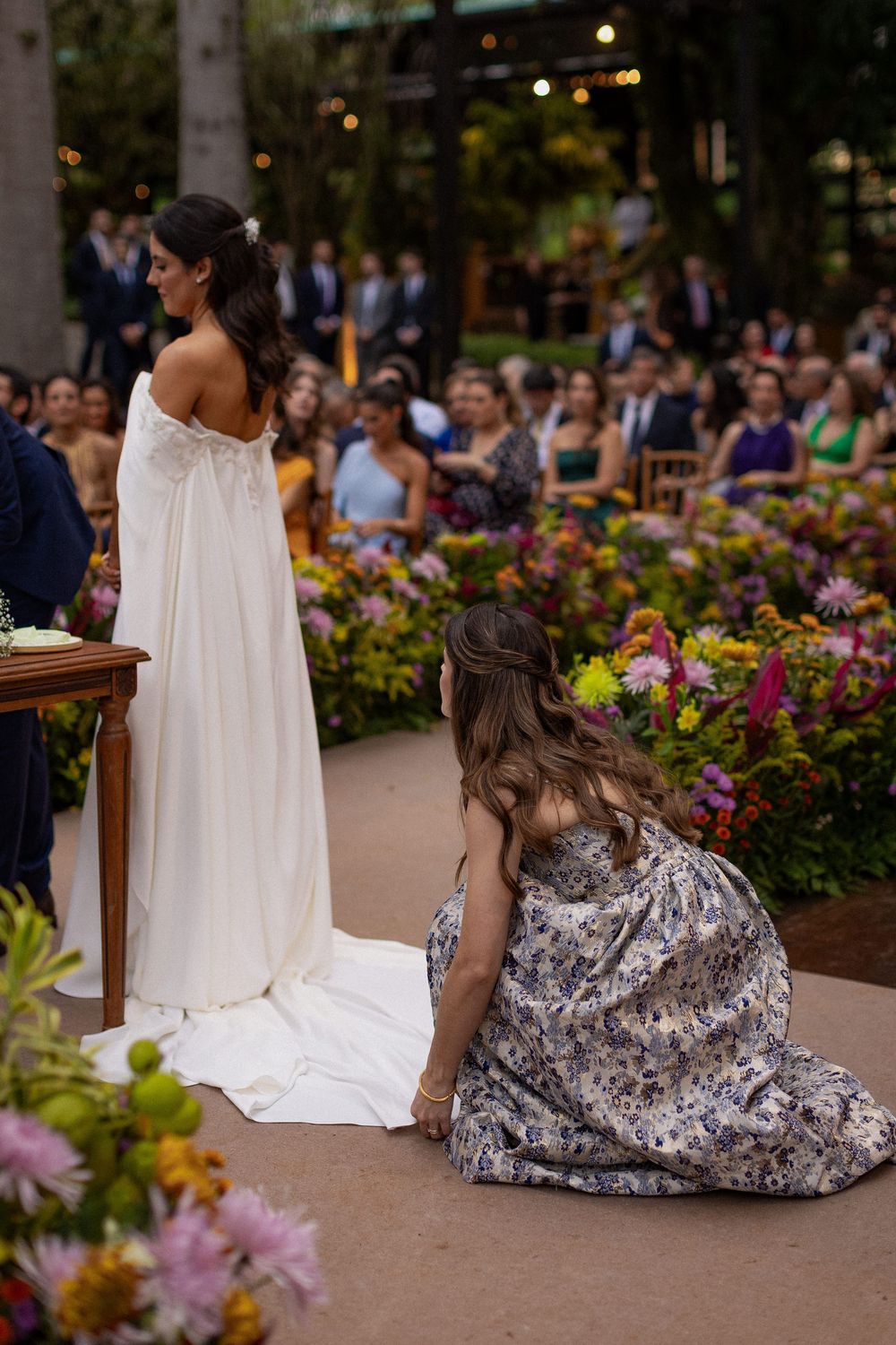 Bride in a flowing white gown stands at an outdoor wedding ceremony while a woman in a floral dress adjusts her train before seated guests.