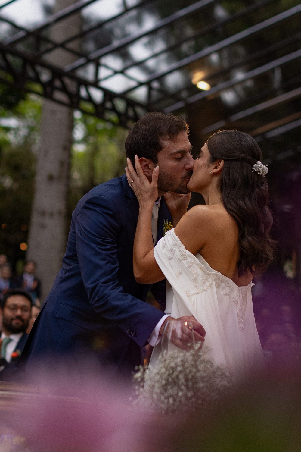 Bride in a white off-the-shoulder dress and groom in a navy suit share a kiss during an outdoor wedding ceremony surrounded by trees.