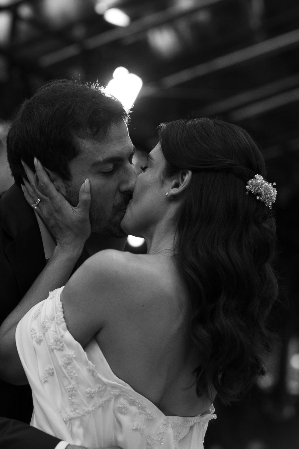 A bride in a lace gown with a floral hair clip shares a romantic kiss with her groom in a black and white wedding photo.