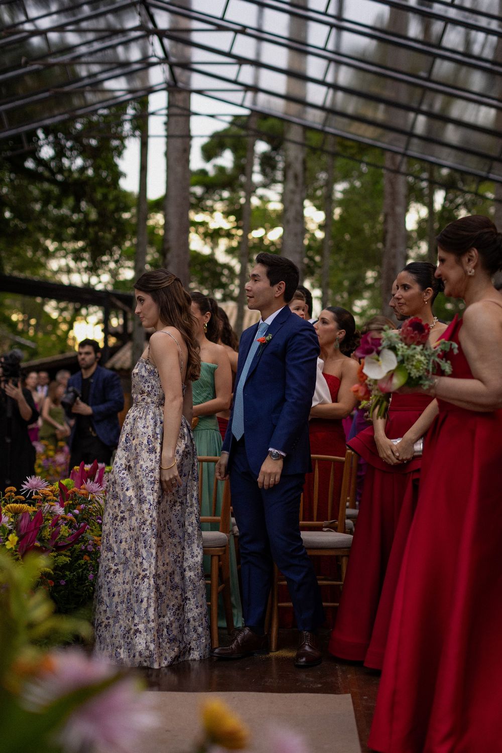 Groom in a blue suit stands at the aisle as bridesmaids in red gowns hold bouquets during an outdoor greenhouse wedding ceremony.