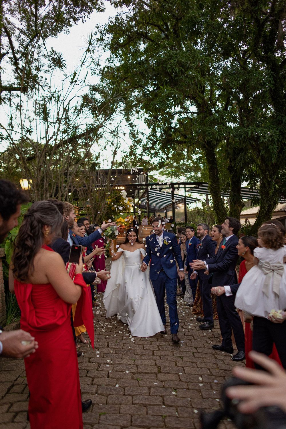 Bride in a white gown and groom in a navy suit walk down a stone path as wedding guests in red dresses and suits cheer under green trees.