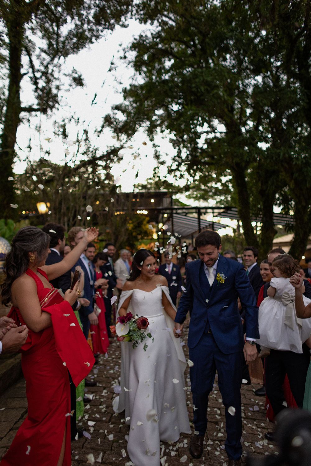 Bride and groom walk hand in hand through a shower of petals as joyful wedding guests cheer under tall trees at an outdoor ceremony.