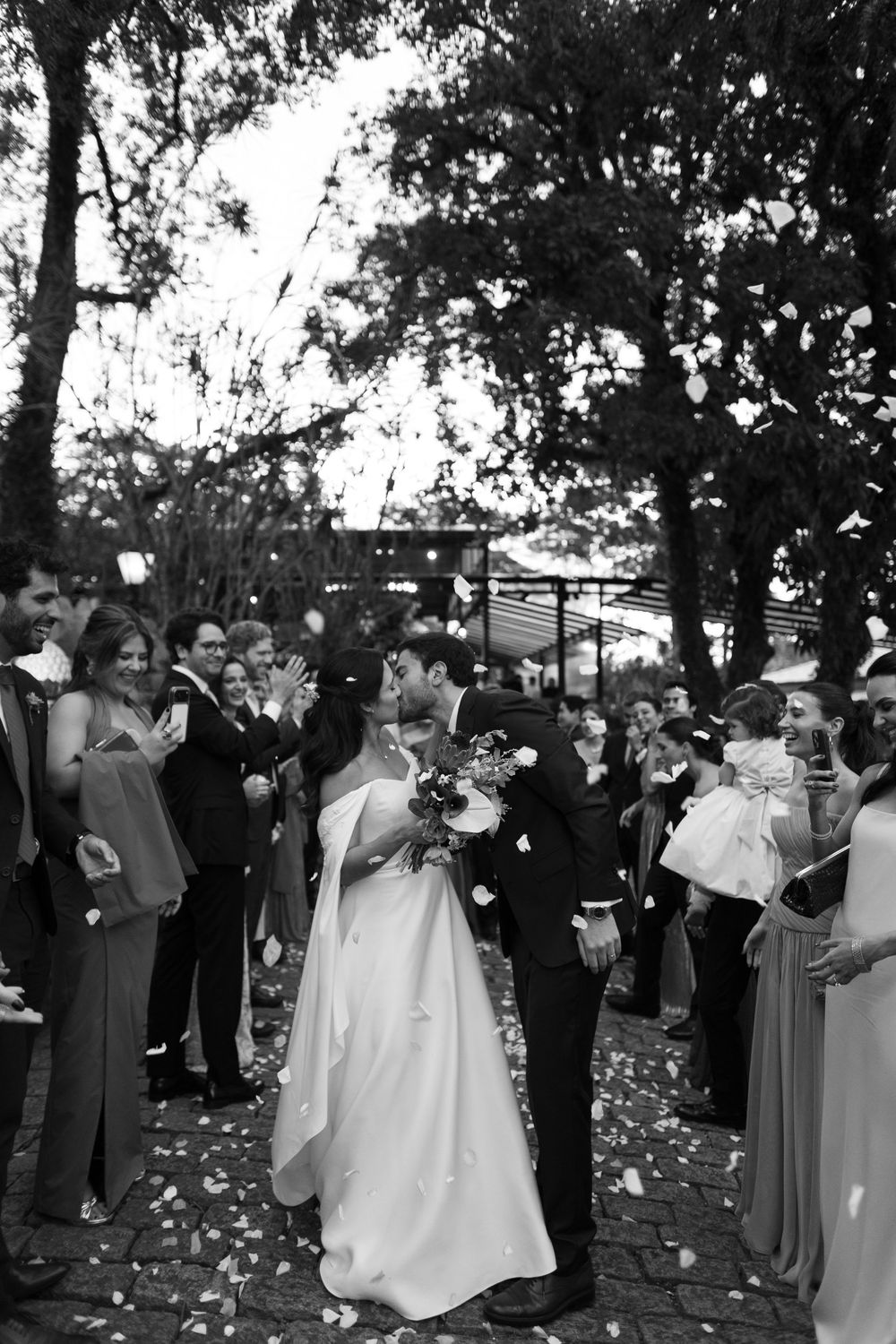 Bride and groom share a kiss as guests cheer and petals fall around them at an outdoor wedding ceremony in black and white.