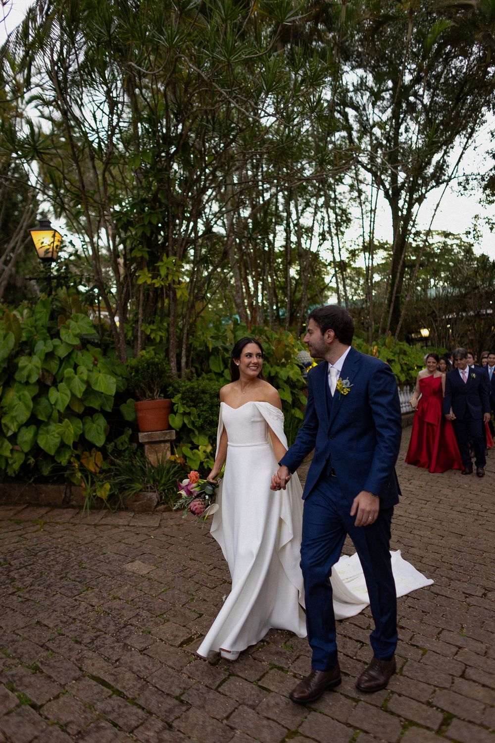 Bride in an off-shoulder white gown and groom in a navy suit walk hand in hand along a brick path in a garden, with guests behind them.