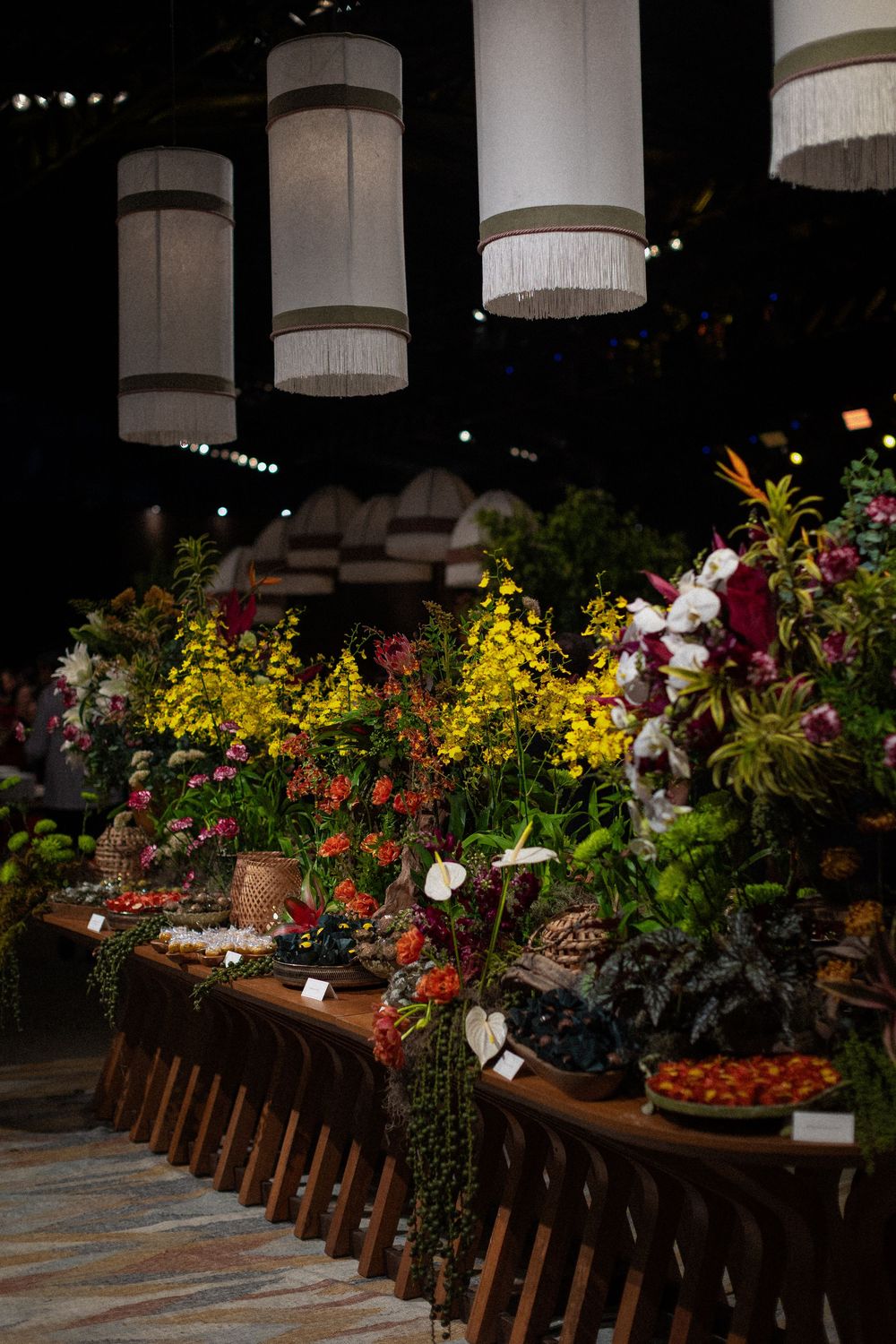 Long wooden display table filled with colorful flower arrangements, yellow orchids and red blooms, beneath fringed white pendant lamps.