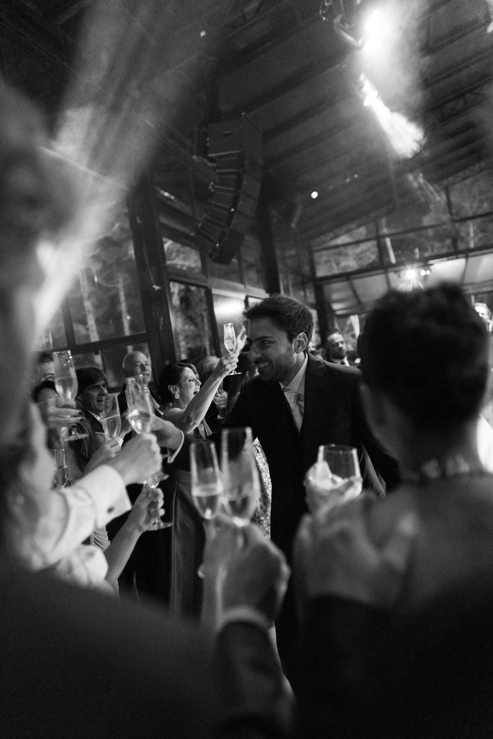Black and white photo of wedding guests raising champagne glasses in a toast as a smiling man in a suit stands in a glass-walled venue.