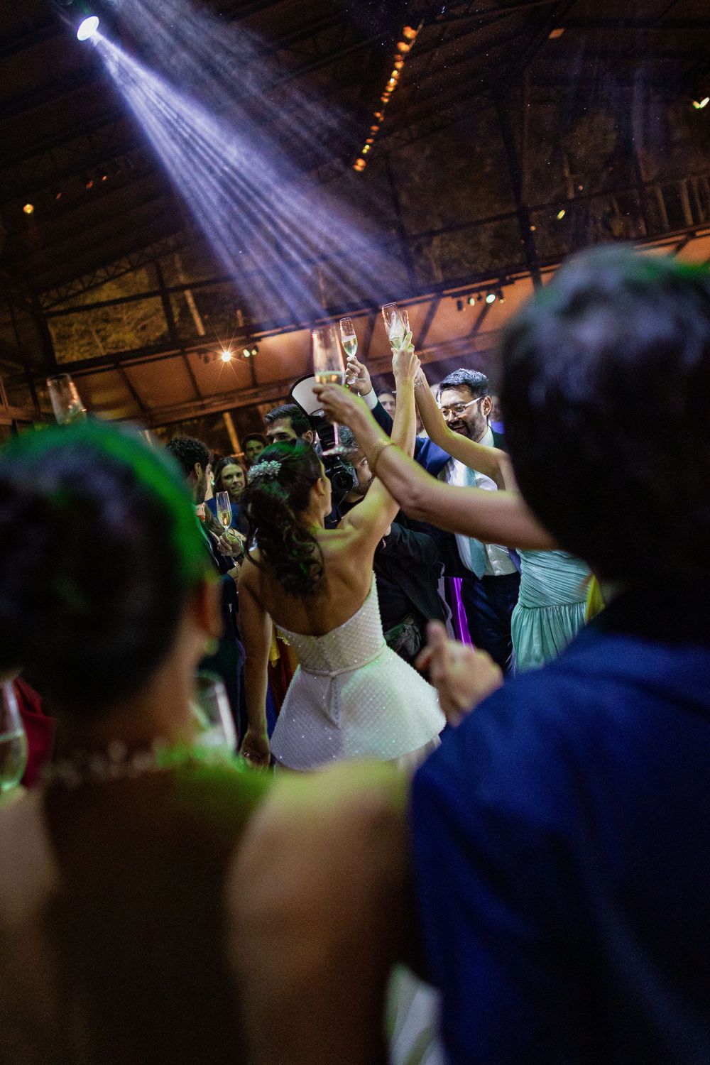 Bride in a white dress raises her arms and a glass in celebration as wedding guests dance under a spotlight at a lively reception.