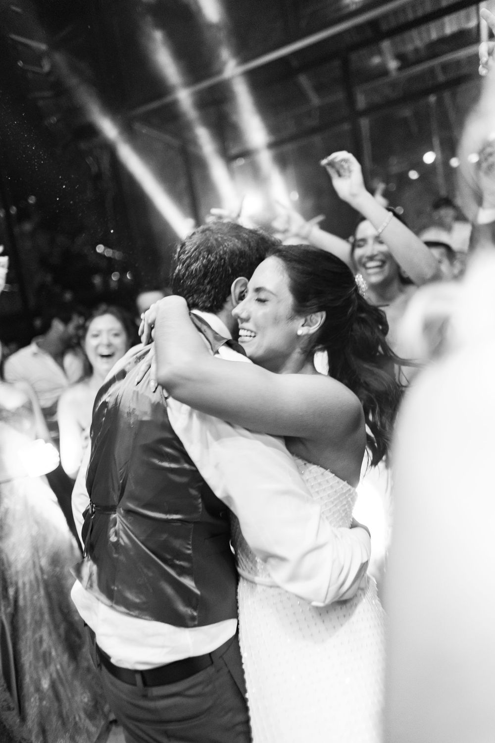 Black and white photo of a smiling bride and groom embracing on a crowded dance floor as guests cheer and celebrate behind them.