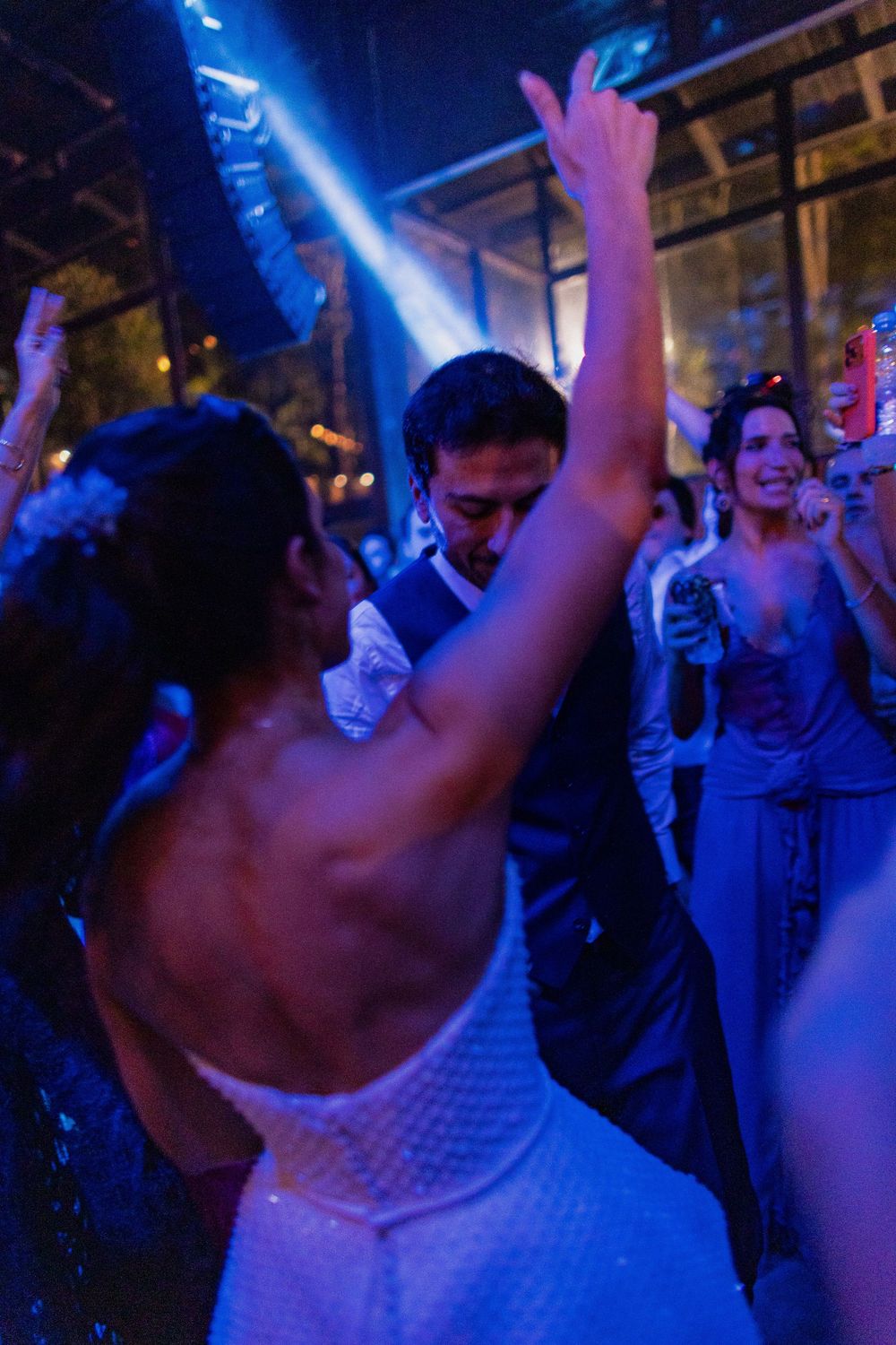 Bride in a sparkling white beaded gown dances with her arm raised as guests celebrate under blue lights at an evening wedding reception.