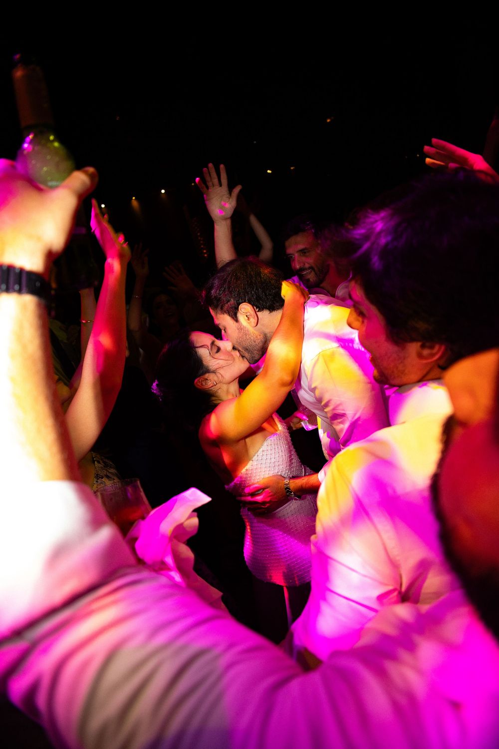 Couple kisses on a crowded dance floor bathed in pink and purple party lighting while guests dance with raised hands around them.
