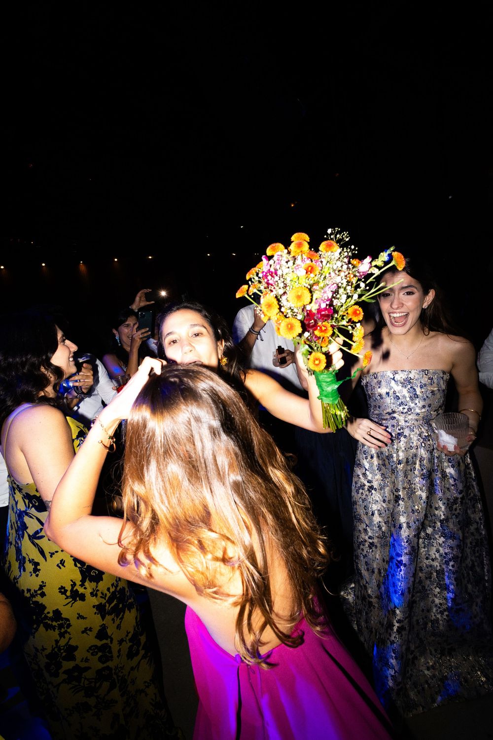 Guests in colorful gowns celebrate at a nighttime wedding as a woman in a floral dress laughs and lifts a bright orange bouquet.