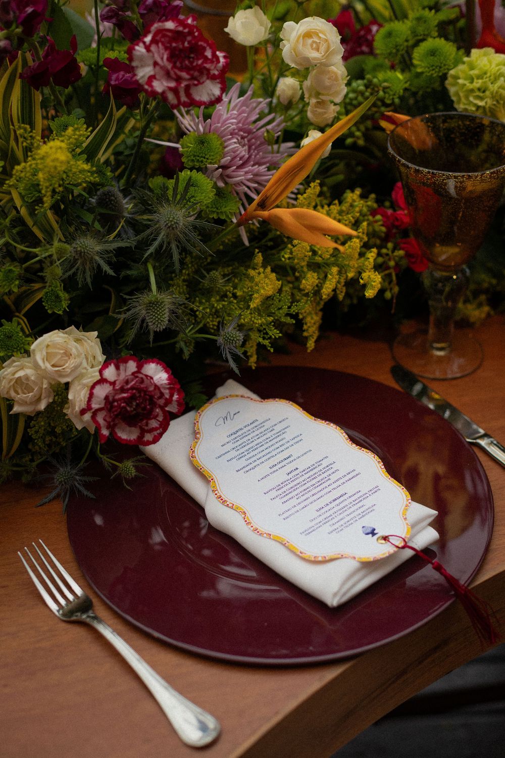 Elegant table setting with a burgundy glass charger, folded napkin with a scalloped menu card, silverware, and colorful floral centerpiece.