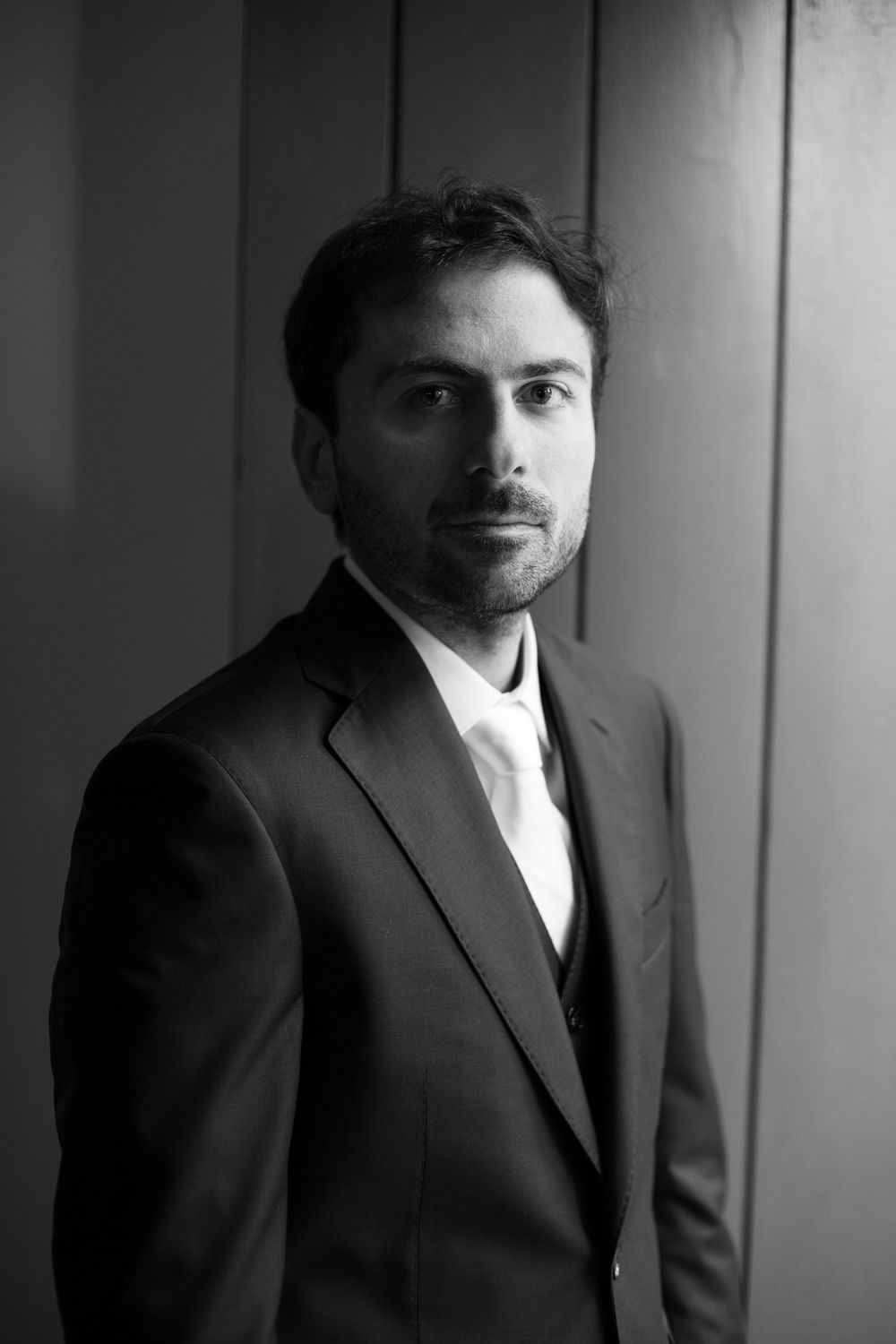 Black and white portrait of a bearded man with tousled dark hair wearing a tailored suit, vest, white shirt and tie.
