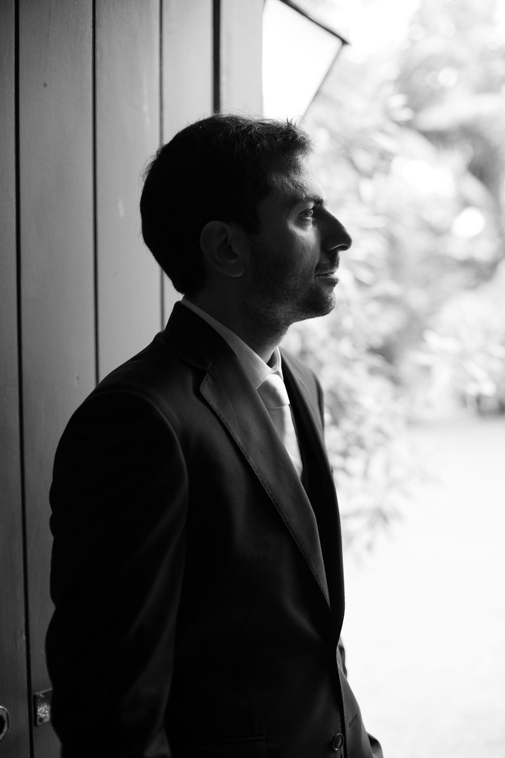 Black and white side profile of a bearded man in a dark suit and tie standing in a wooden doorway, gazing out toward blurred greenery.