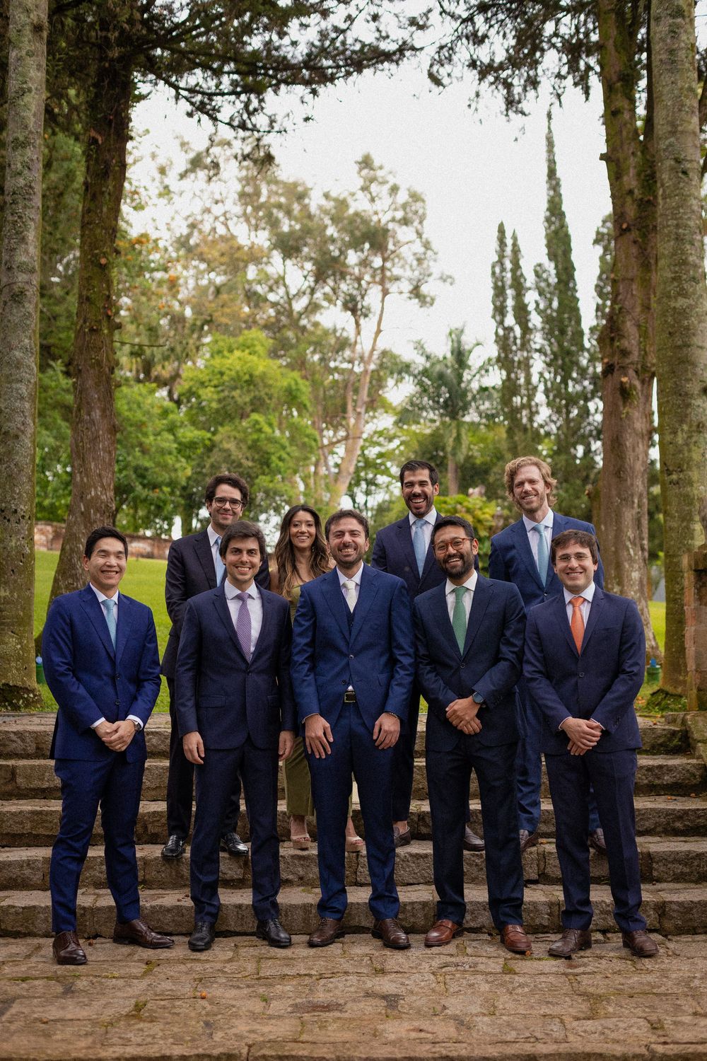 Group of men in navy suits and ties posing together on stone steps in a lush green park with tall trees.