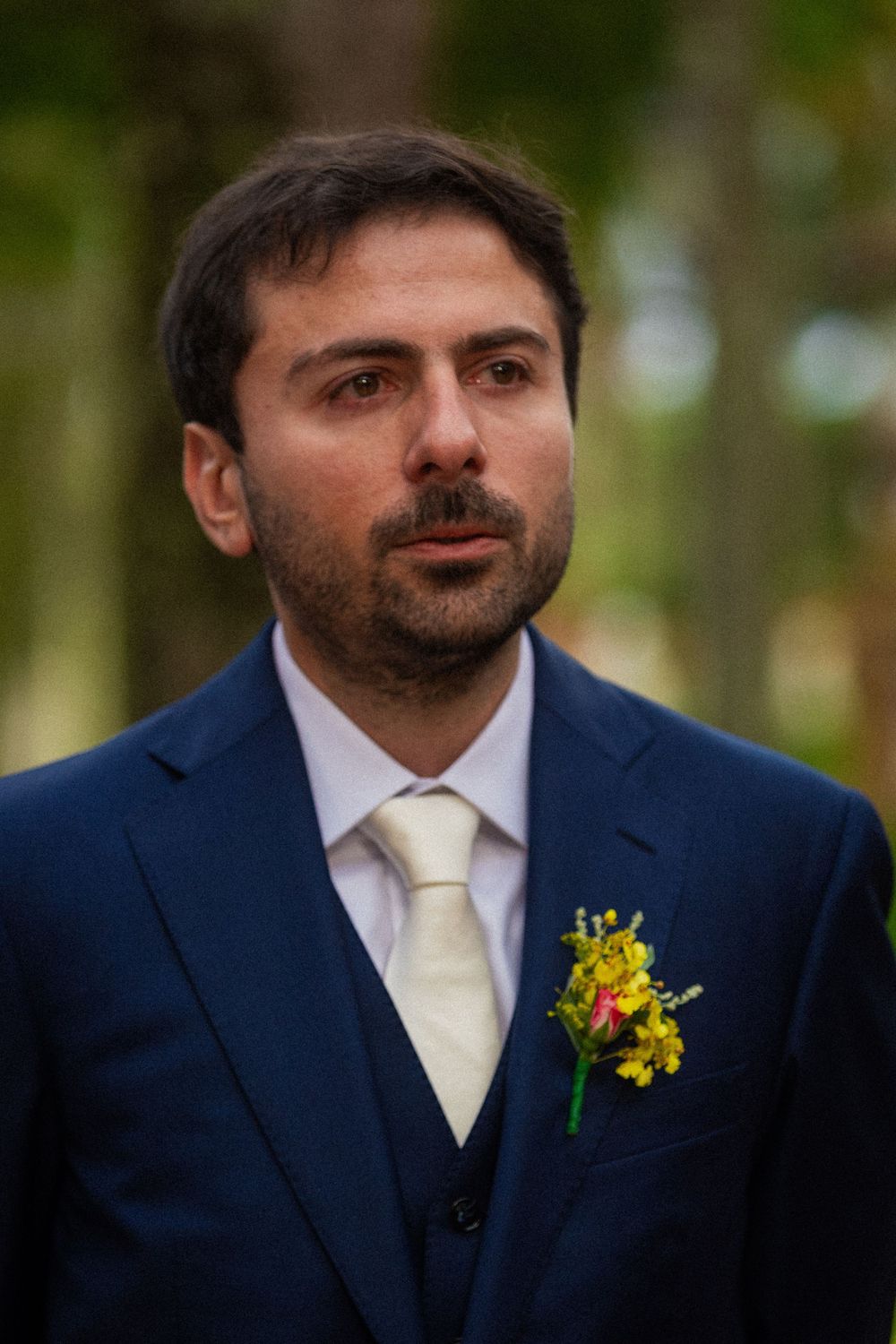 Groom in a navy blue suit, vest and cream tie wearing a yellow flower boutonniere, standing outdoors among blurred trees.