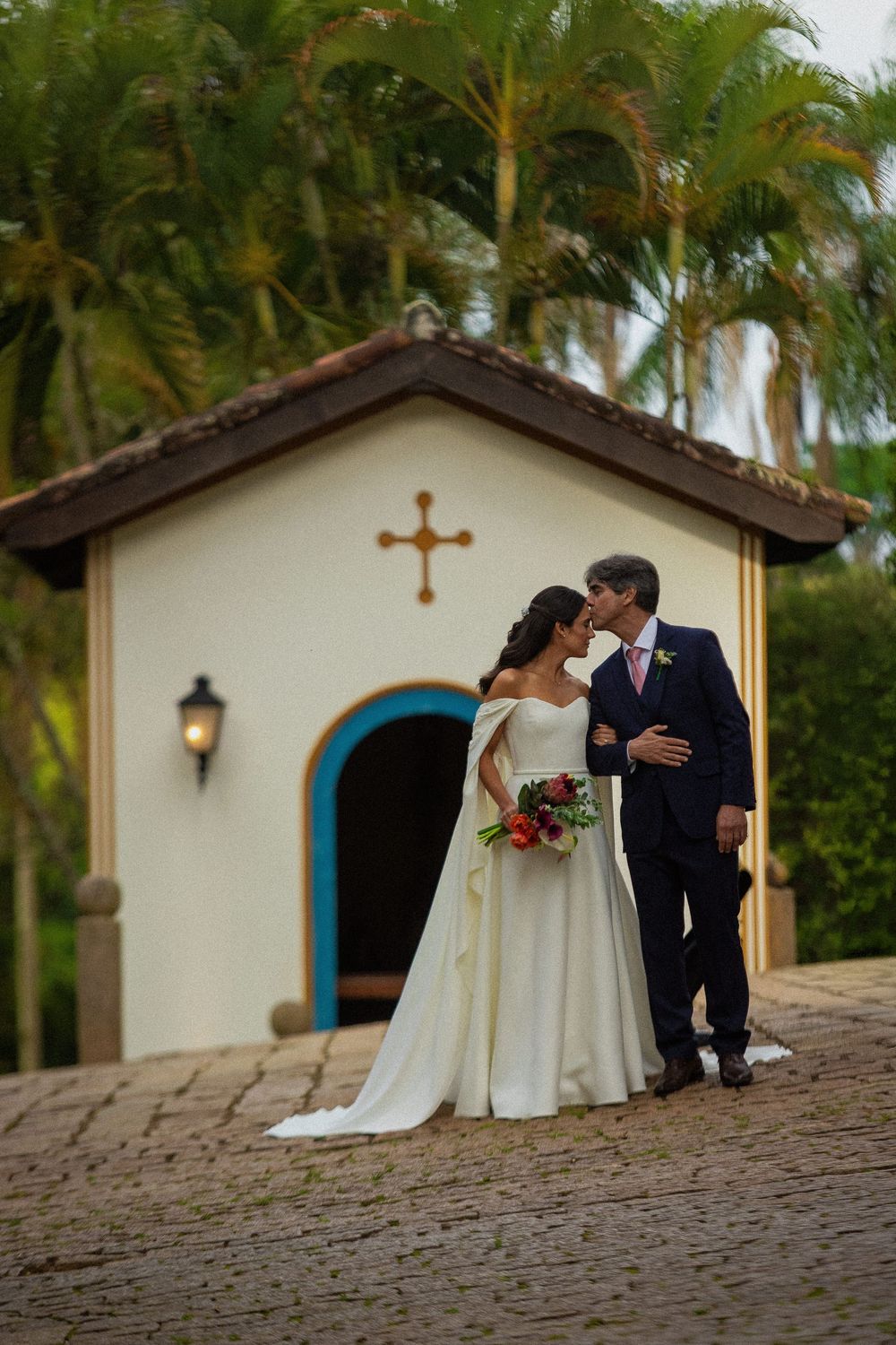 Bride in a flowing white gown and groom in a navy suit kiss in front of a small white chapel with a blue arched door and palm trees.