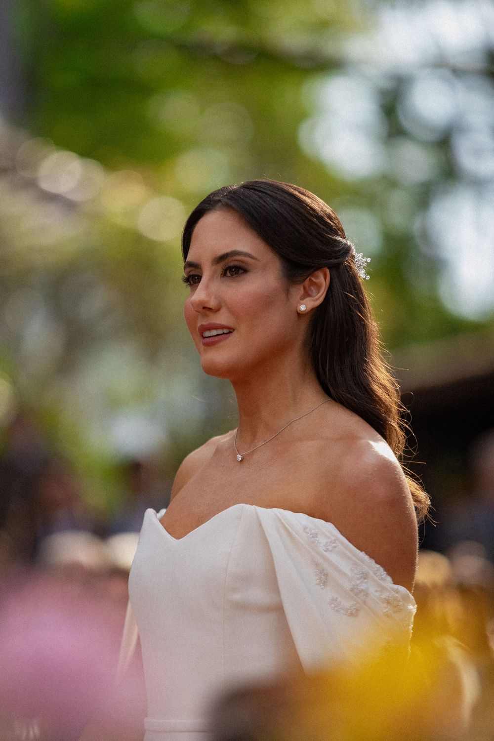 Smiling bride with dark hair pinned back, wearing an off-the-shoulder white gown and pendant necklace, in a softly blurred garden setting.