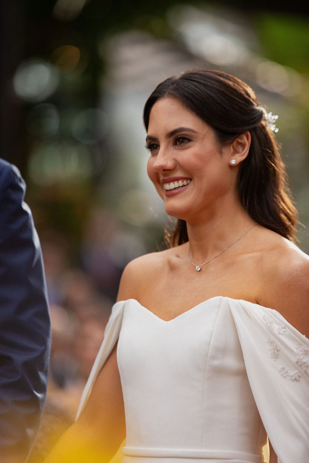 Smiling bride in an off-the-shoulder white wedding gown with cape sleeves and a delicate necklace at an outdoor ceremony.